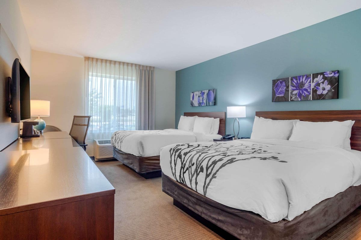 Quarto de hotel com duas camas, uma escrivaninha, televisão de tela plana, abajur e obras de arte nas paredes.
