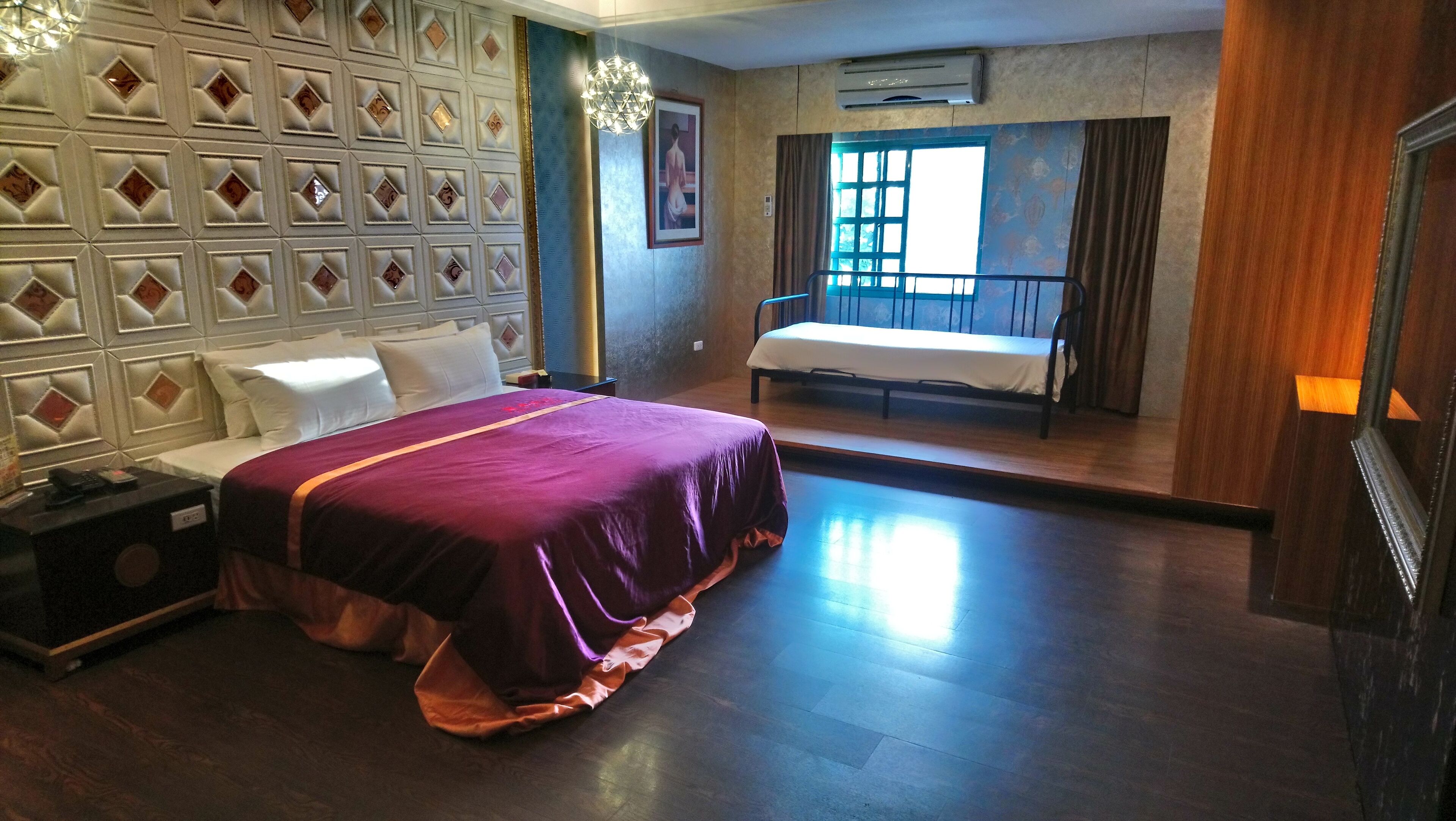 Una habitación de hotel con una cama grande, una mesita de noche con lámpara, una pequeña mesa con una silla, una ventana con cortinas y una pared con un diseño geométrico.