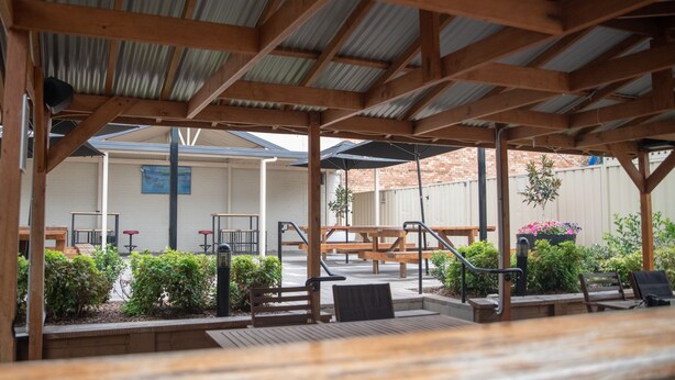 Un espace extérieur aménagé en terrasse, avec des bancs et des tables en bois, entouré de plantes et surmonté d’un toit en métal ondulé.