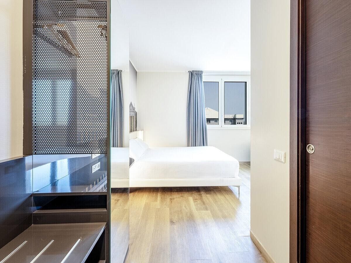 A hotel room with a bed, a window with curtains, a door, and a view of the city.