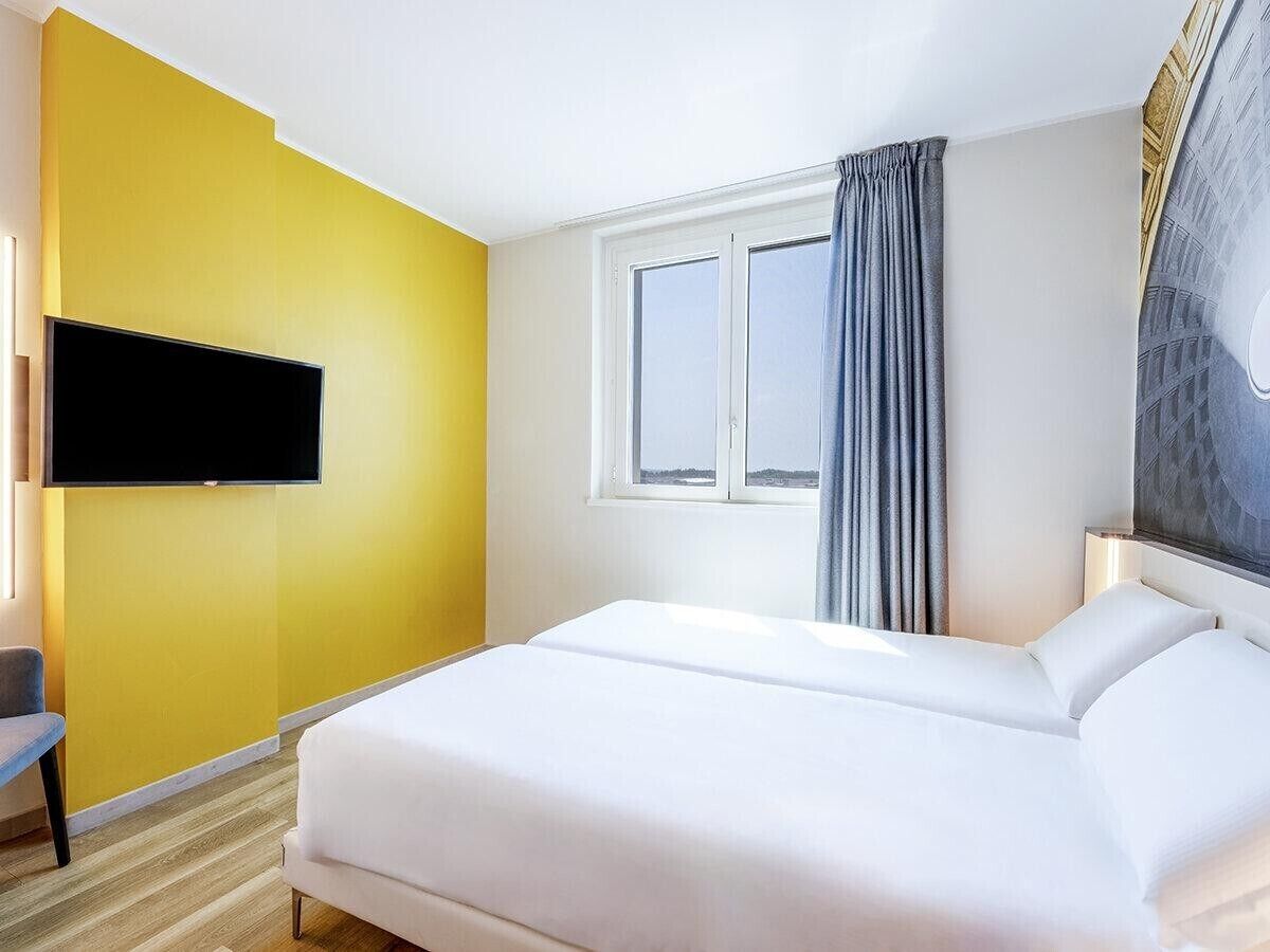 Ein Hotelzimmer mit einem Bett, einem Flachbildfernseher, einem Fenster mit Vorhängen und einer Wand mit Wandgemälde.