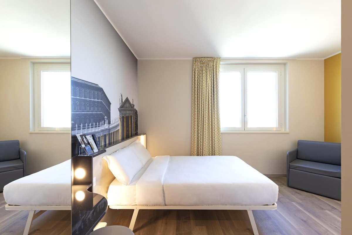 Una habitación moderna con una cama, dos sofás grises, una ventana con cortinas y un televisor fijado a la pared.