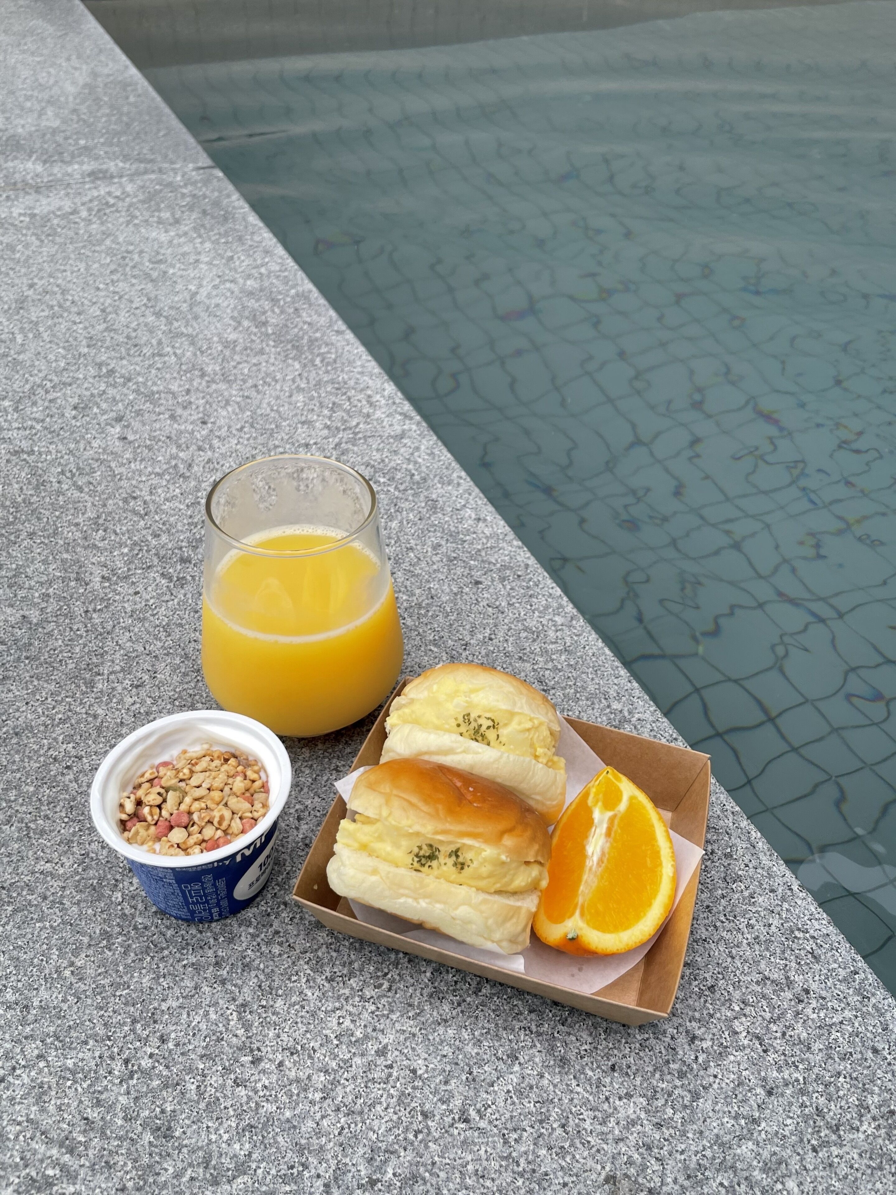 A breakfast setup by a poolside with a glass of orange juice, a bowl of cereal, and two pastries with an orange slice.