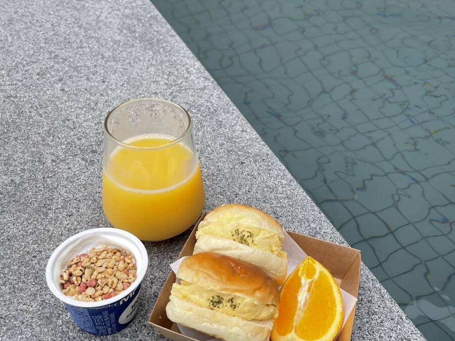 A breakfast setup by a poolside with a glass of orange juice, a bowl of cereal, and two pastries with an orange slice.