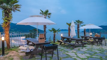 An outdoor seating area with wooden tables and benches, surrounded by palm trees and umbrellas, overlooking a body of water and mountains.