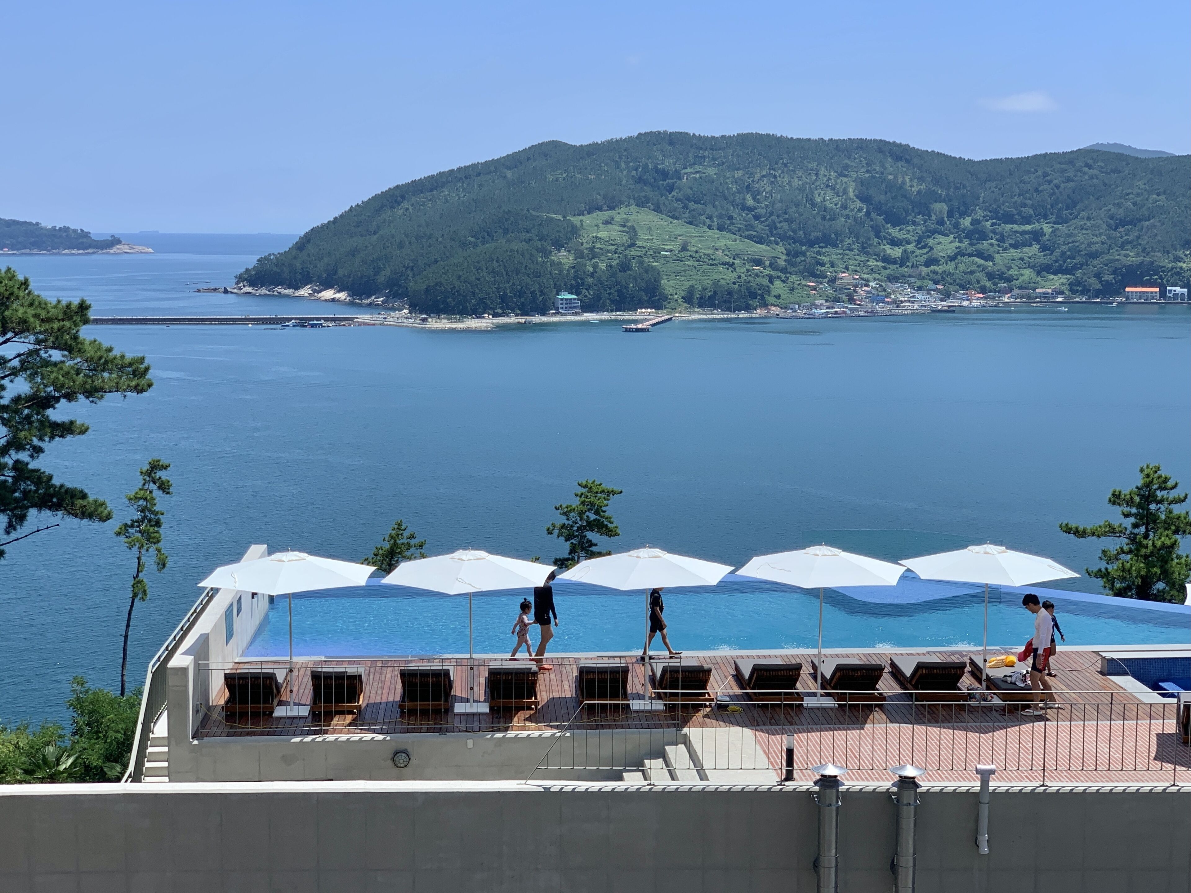 A rooftop pool with umbrellas, overlooking a body of water and a forested hill.