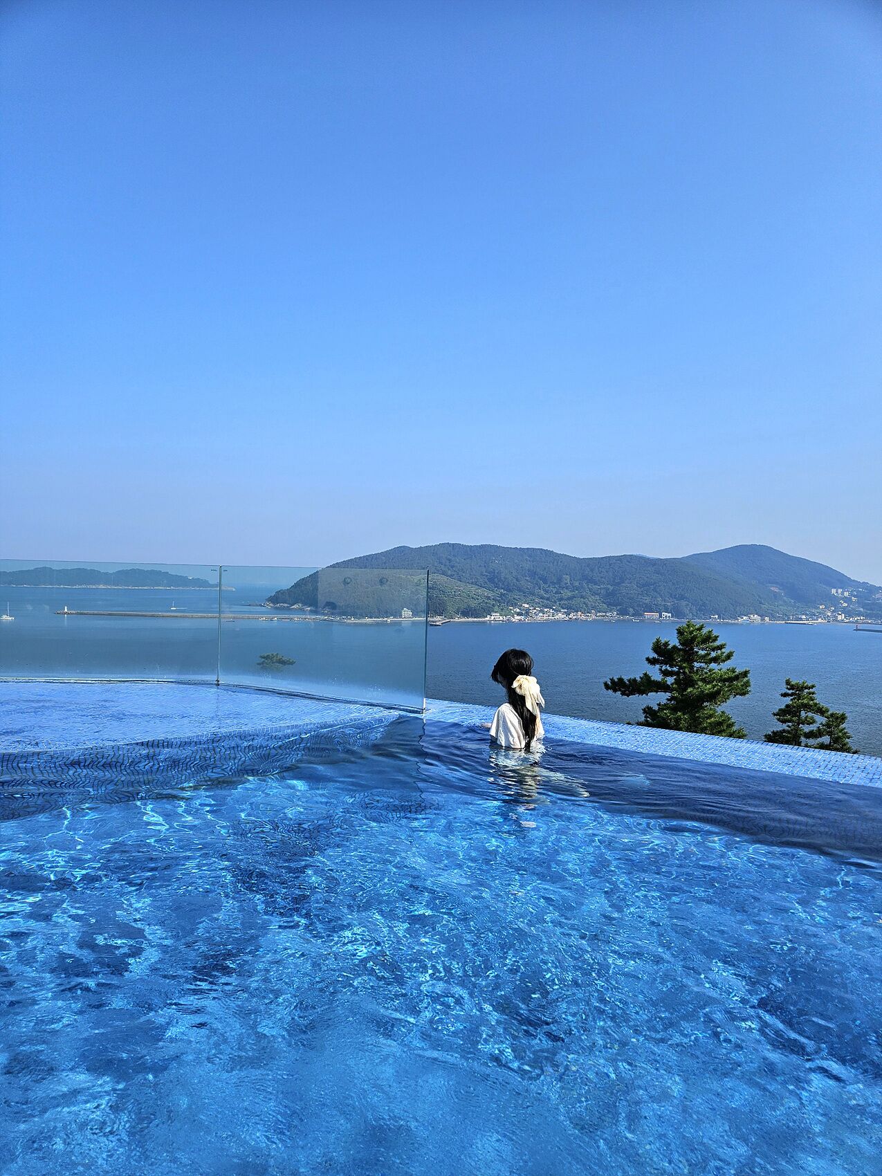 A person standing in a clear blue infinity pool with a view of a distant shoreline and hills.