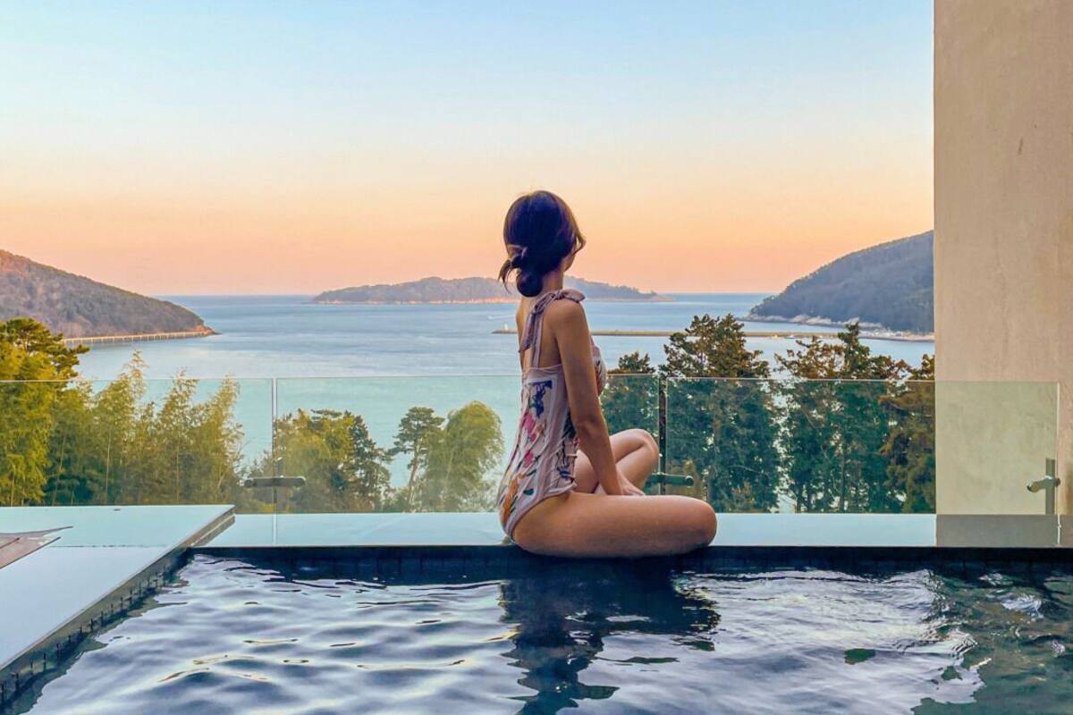 A person sitting by a pool with a view of the sea and hills.