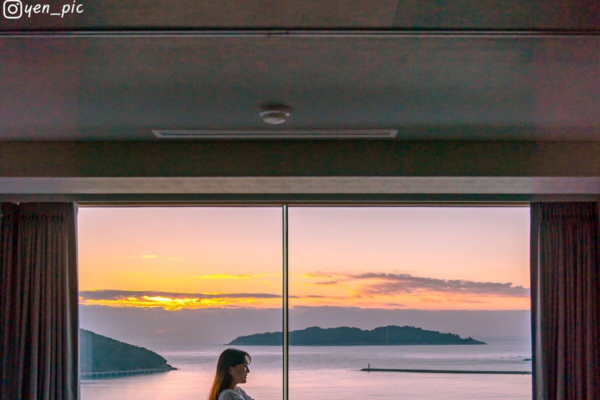 A room with a large window offering a view of the sea and distant land during sunset.