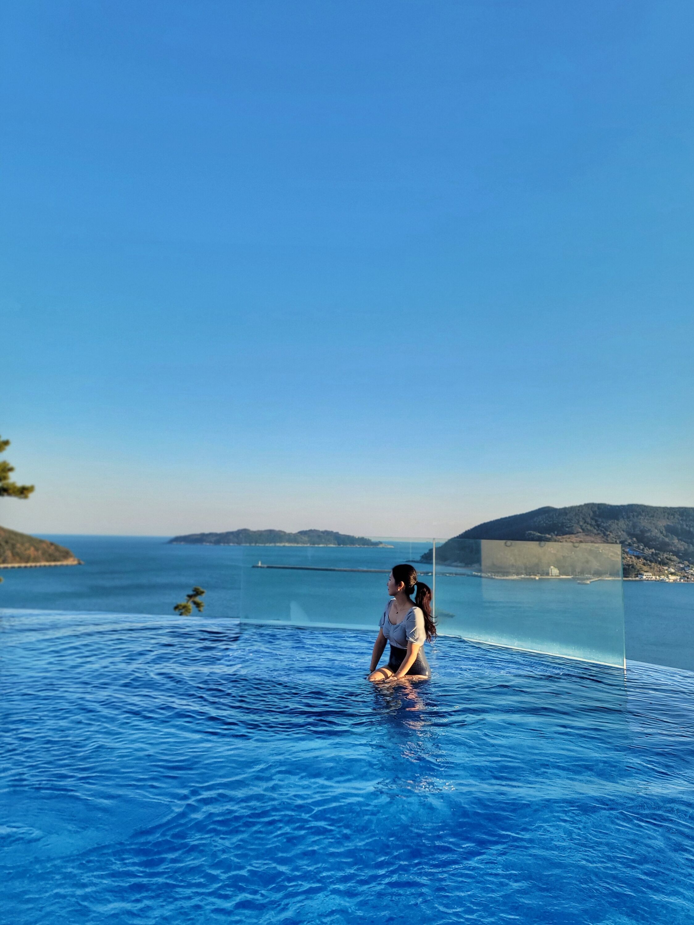 A person sitting in a clear-bottomed infinity pool with a view of the sea and distant hills.