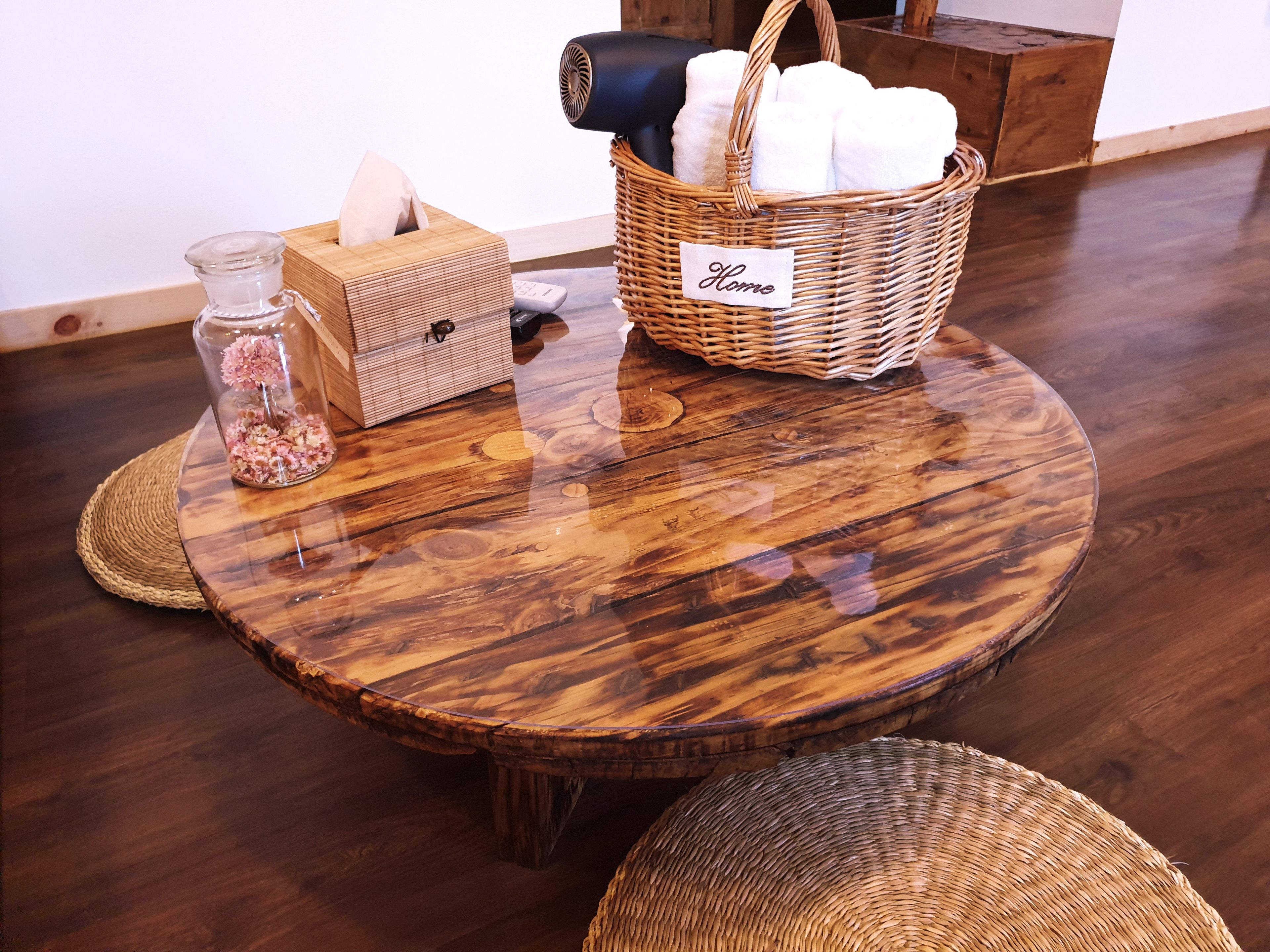A round wooden table with a woven placemat, a glass vase with pink flowers, a tissue box, a basket labeled "Home," and a remote control.