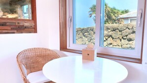 Quarto com uma cadeira de vime, uma mesa branca redonda e uma janela com vista para uma parede de pedra e palmeiras.
