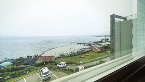 A coastal view from a window overlooking a beach with a parked car and buildings in the background. - Fantastic Ocean View Sydney Hotel (Jeju City)