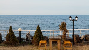 A coastal scene with a wooden bench, two chairs, and a table by the water, with a railing and a lamp post. - Fantastic Ocean View Sydney Hotel (Jeju City)