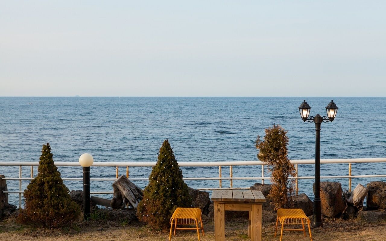 A seaside view with a railing, two chairs, a small table, and two street lamps.
