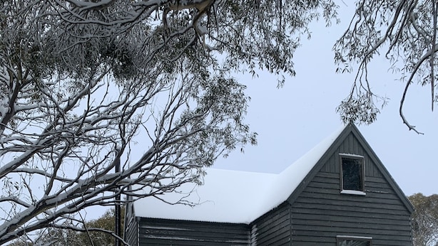 Une cabane dont le toit est recouvert de neige, avec un porche à l’avant et une table de pique-nique.