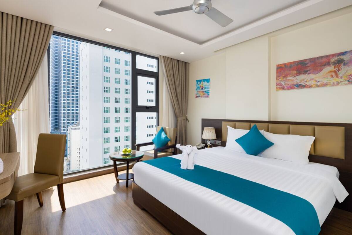 Une chambre d’hôtel avec un grand lit, un bureau, une chaise, un ventilateur de plafond et une vue sur de hauts immeubles.