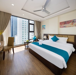 A hotel room with a large bed, a desk, a chair, a ceiling fan, and a view of tall buildings.