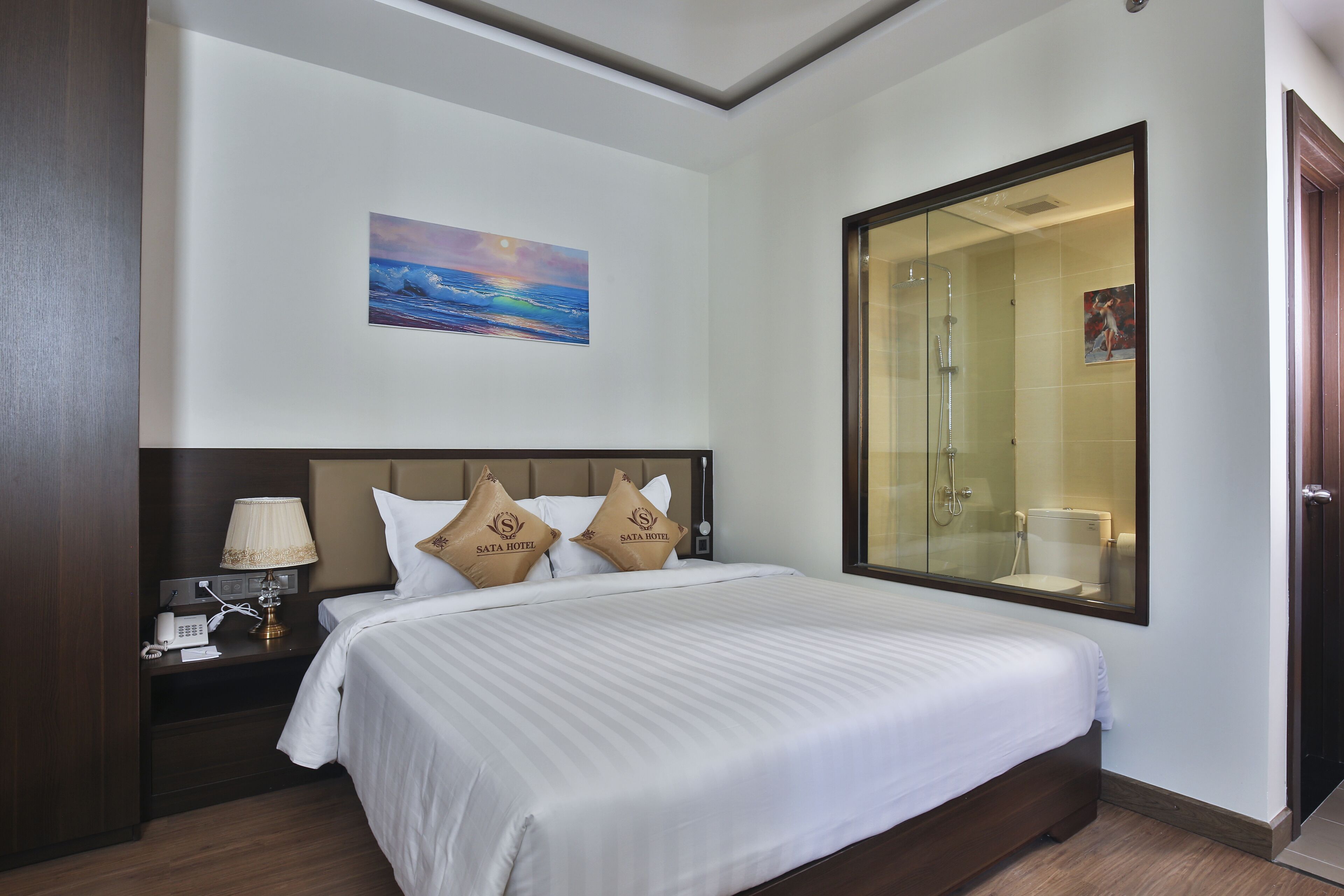 A hotel room with a large bed, a nightstand with a lamp, a painting on the wall, and a glass-enclosed shower area.