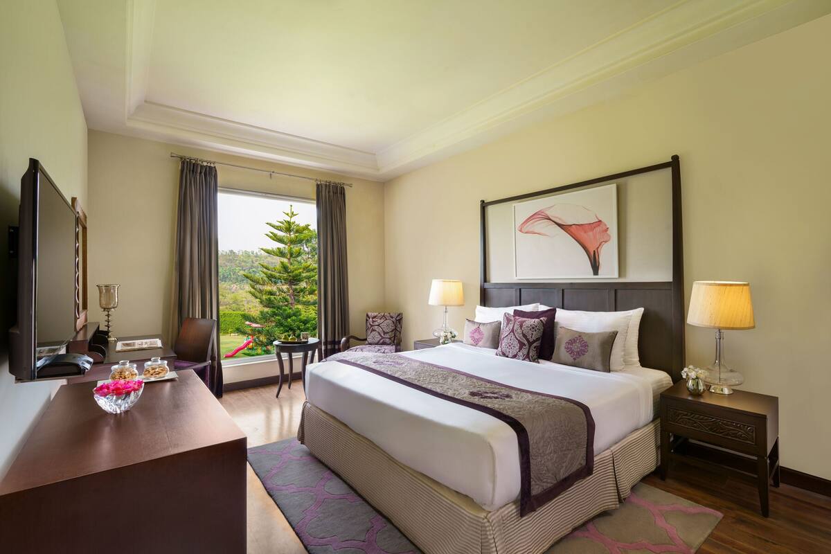 Habitación de hotel con una cama grande, un escritorio, una silla, una mesita de noche y una ventana con vistas a zonas verdes.