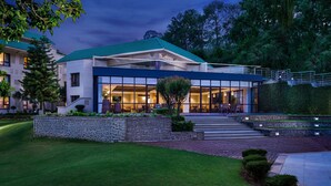 A modern building with a glass facade, a stone retaining wall, and a green roof. - Vivanta Katra Vaishno Devi (Reasi)