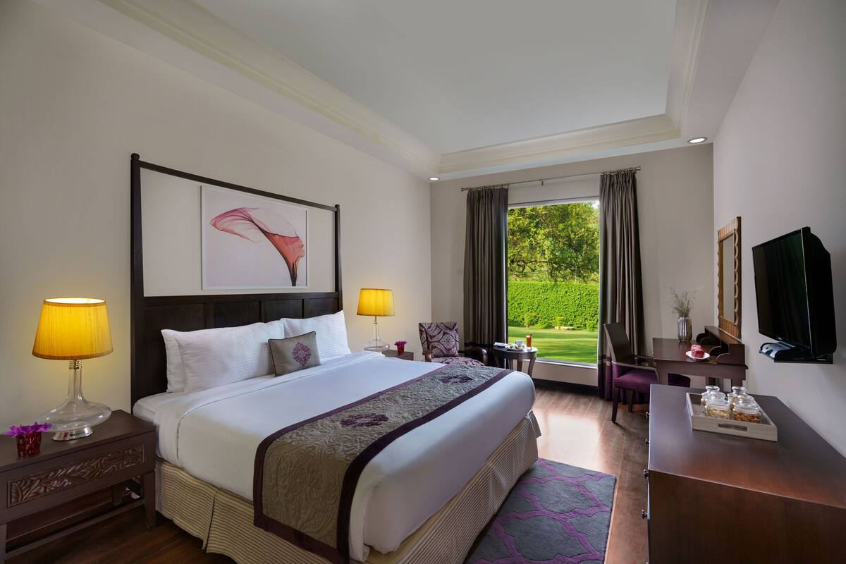 Habitación de hotel con una cama grande, dos lámparas de noche, un escritorio con una silla, un televisor y una ventana con vistas a zonas verdes.