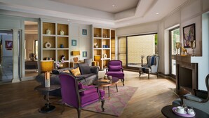 A spacious living room with a fireplace, a built-in bookshelf, and a large window offering a view of the outside. - Vivanta Katra Vaishno Devi (Reasi)