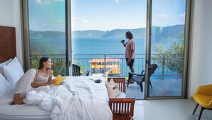 A bedroom with a large window overlooking a lake, a balcony with a chair, and a bed with a pillow and a book. - Equinoccio Hotel (Santa Ana)
