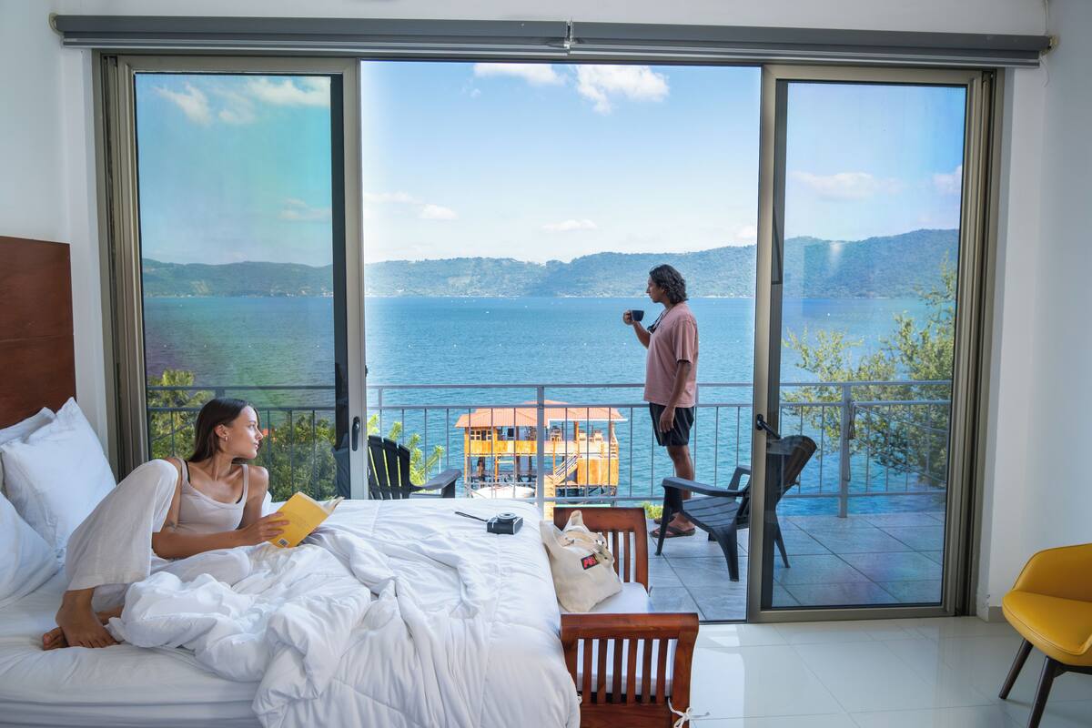 A bedroom with a large window overlooking a lake, a balcony with a chair, and a bed with a pillow and a book.