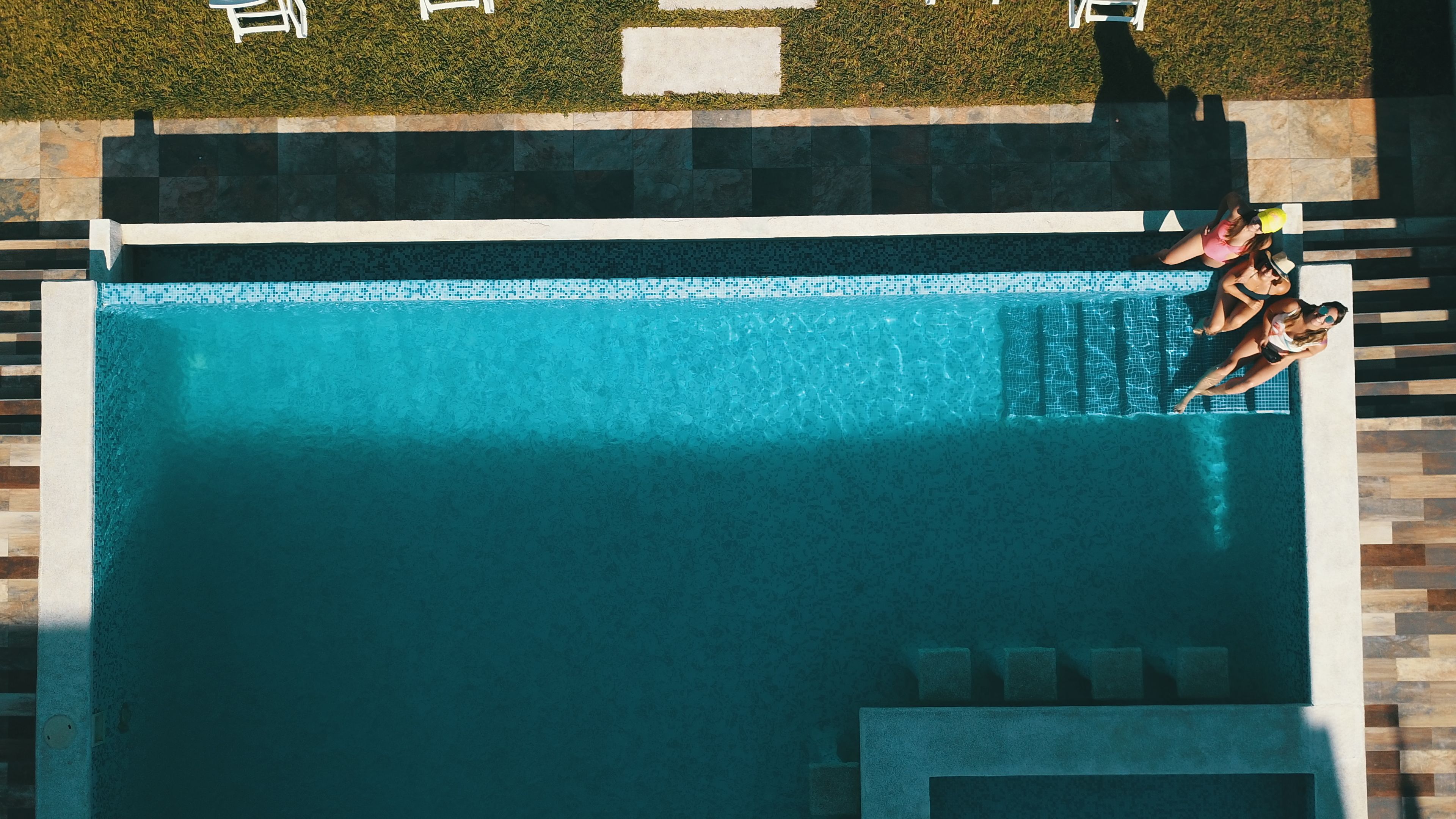 Vue aérienne d’une piscine rectangulaire dont le bord est recouvert de carreaux, entourée d’une bordure en pierre et d’une zone herbeuse.