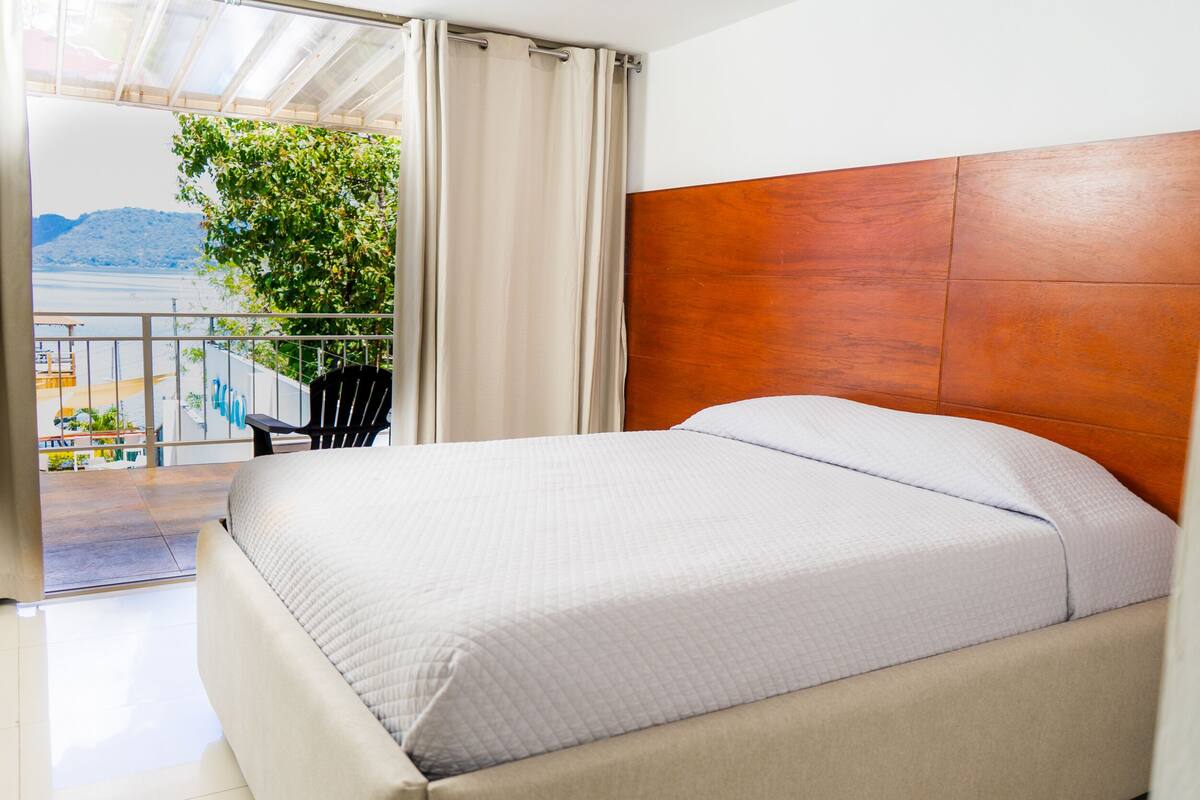 A bedroom with a bed, a wooden headboard, a balcony with a view of the sea, and a chair.