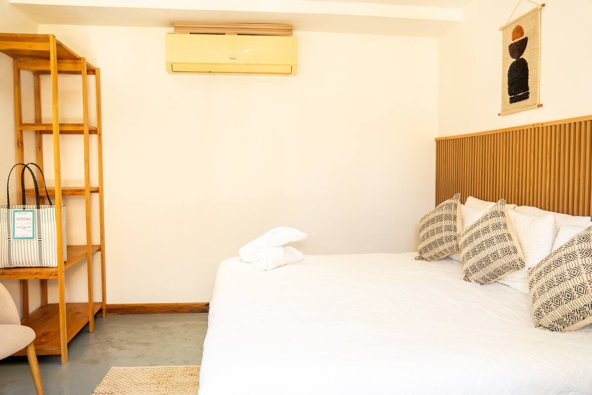 A bedroom with a bed, pillows, a wooden shelf, a hanging air conditioner, and a wall-mounted artwork.