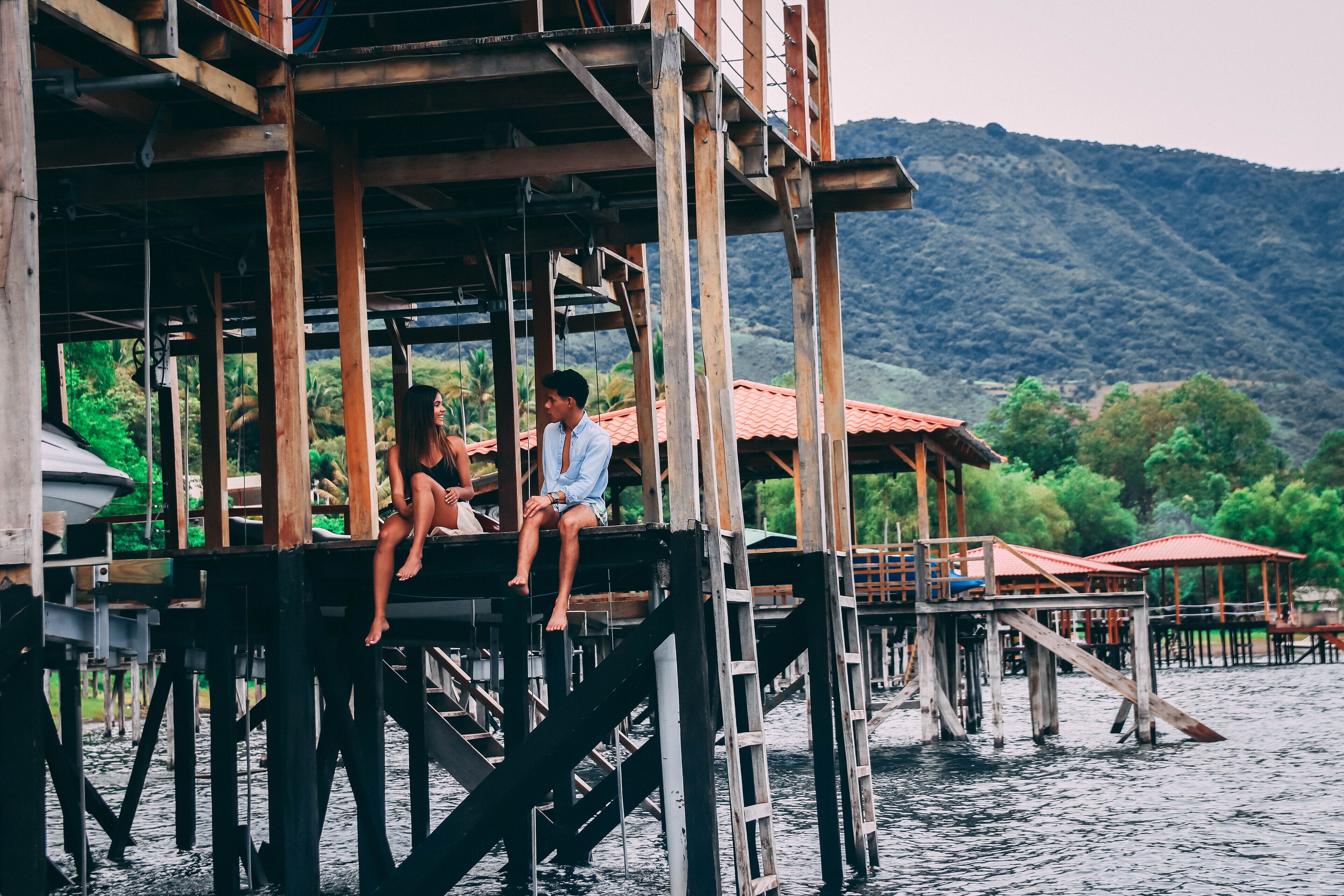 A wooden structure with a platform extending over water, featuring a person sitting and another standing.