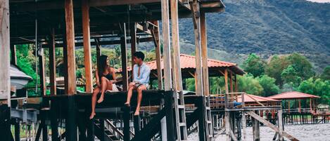 A wooden structure with a platform extending over water, featuring a person sitting and another standing.
