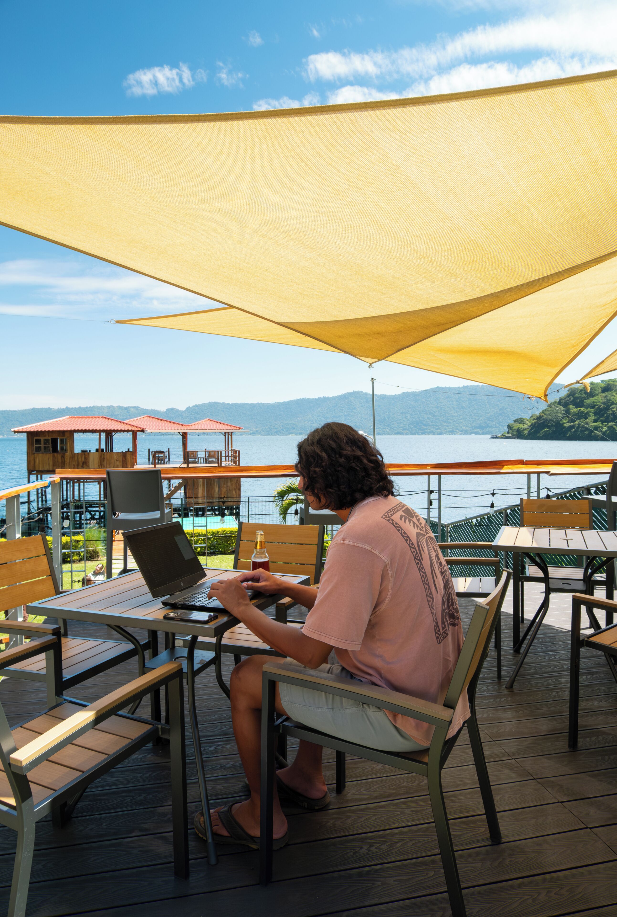 A person using a laptop at a wooden table with a view of a lake and buildings in the background.
