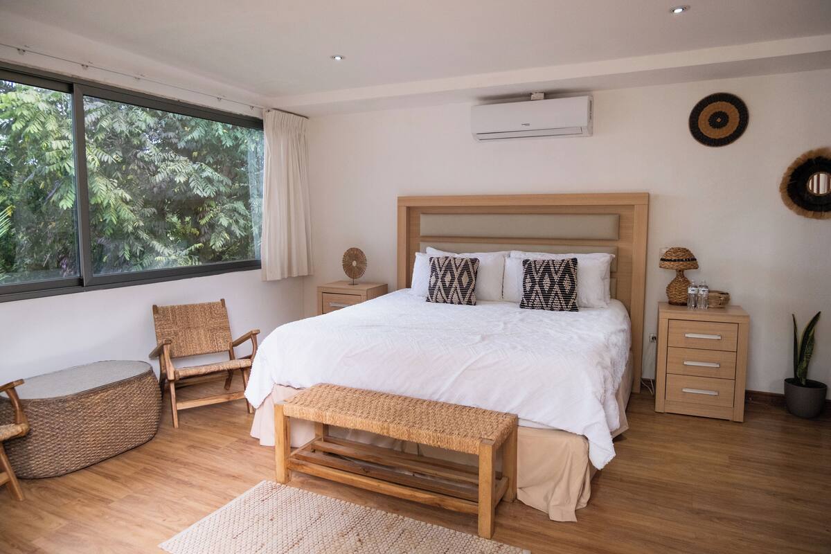 A modern bedroom with a large bed, wooden furniture, a window with a view of trees, and a small bedside table with a lamp.