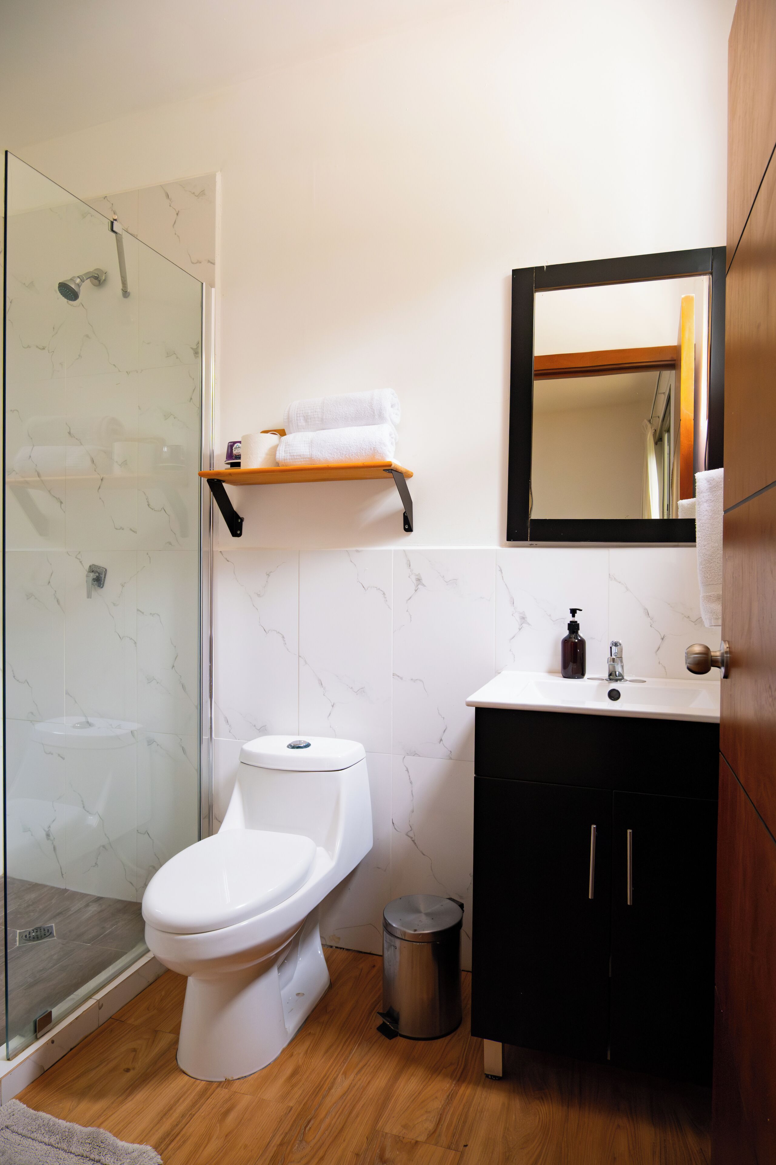 A modern bathroom with a glass-enclosed shower, a white toilet, a black cabinet, and a wooden shelf with towels.