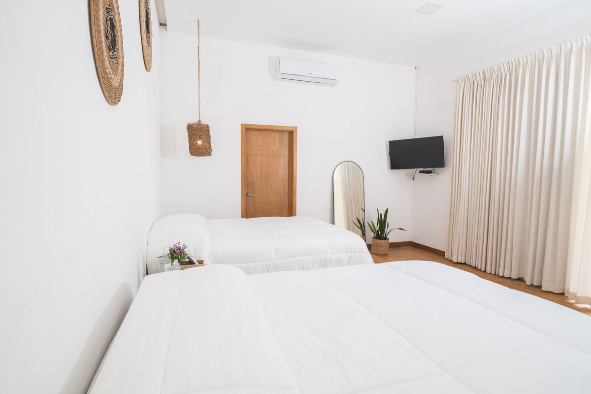 A hotel room with two beds, a wooden door, a flat-screen TV, a hanging light fixture, and a large window with curtains.