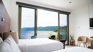 Habitación de hotel con dos camas, un balcón con vistas al mar, un televisor montado en la pared y una pequeña mesa con una planta.