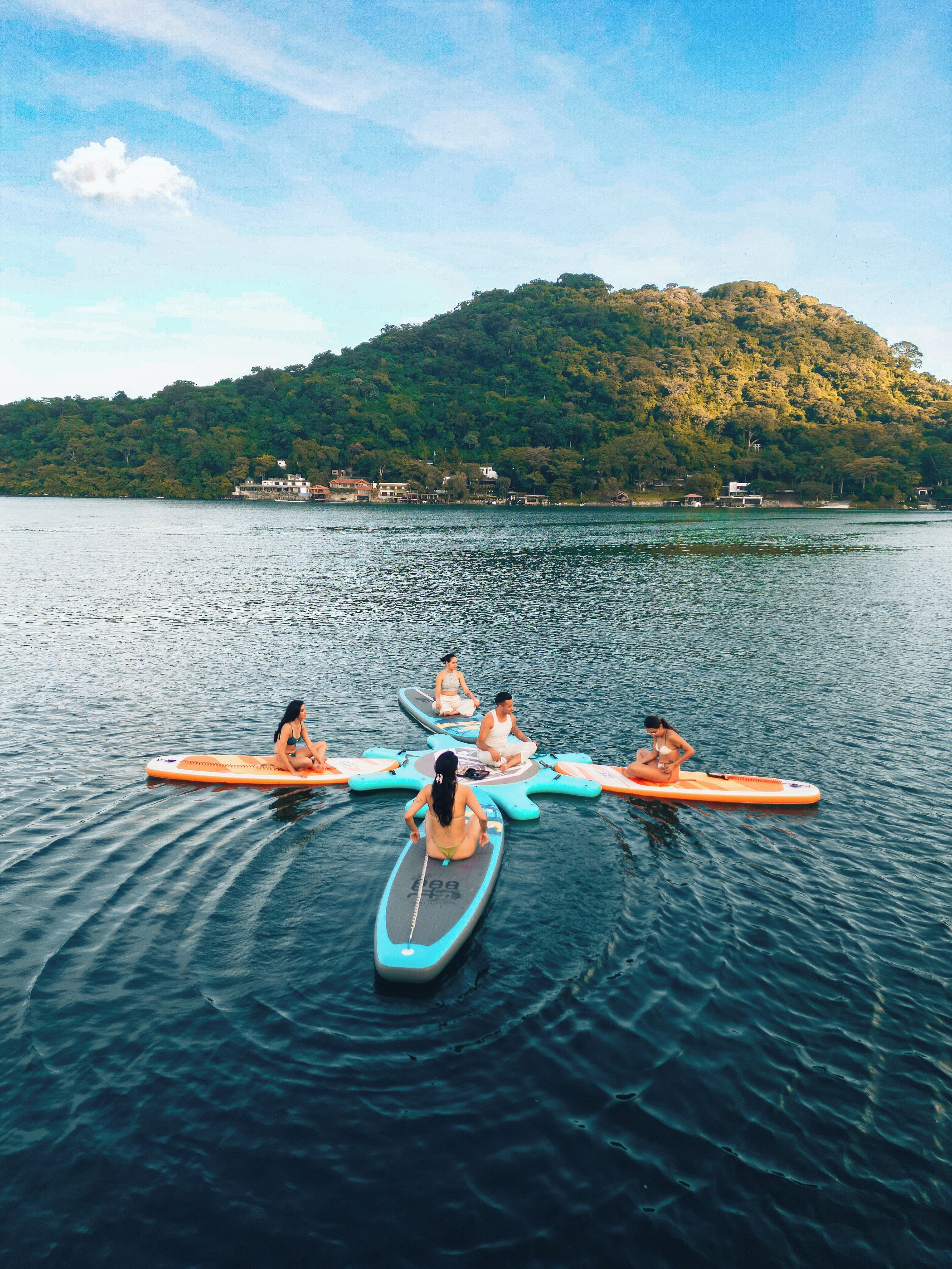 Eine Gruppe von Menschen sitzt auf Paddleboards auf einem ruhigen See, im Hintergrund ist ein bewaldeter Hügel zu sehen.