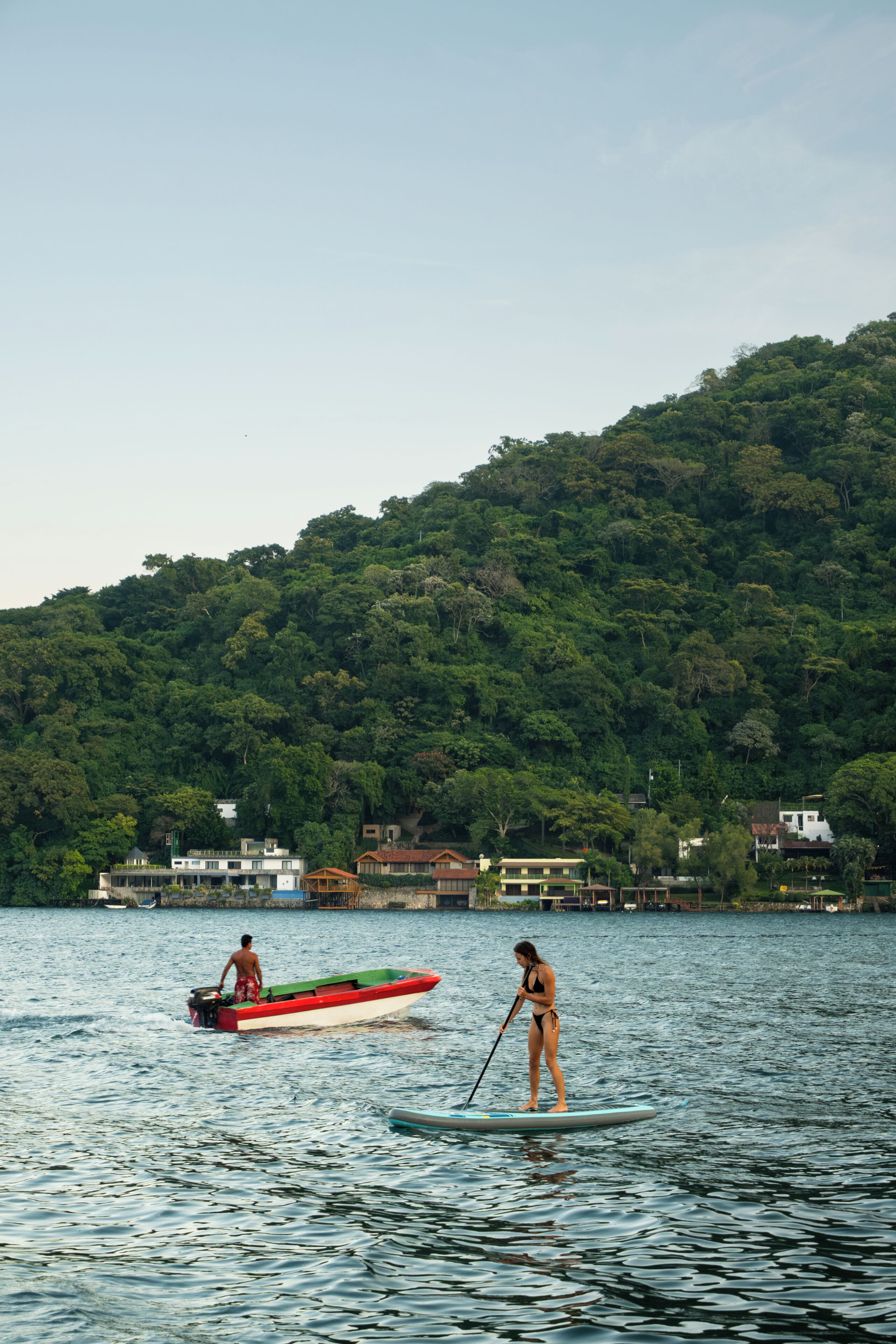 A lake with paddle boats, a forested hill in the background, and buildings along the shore.