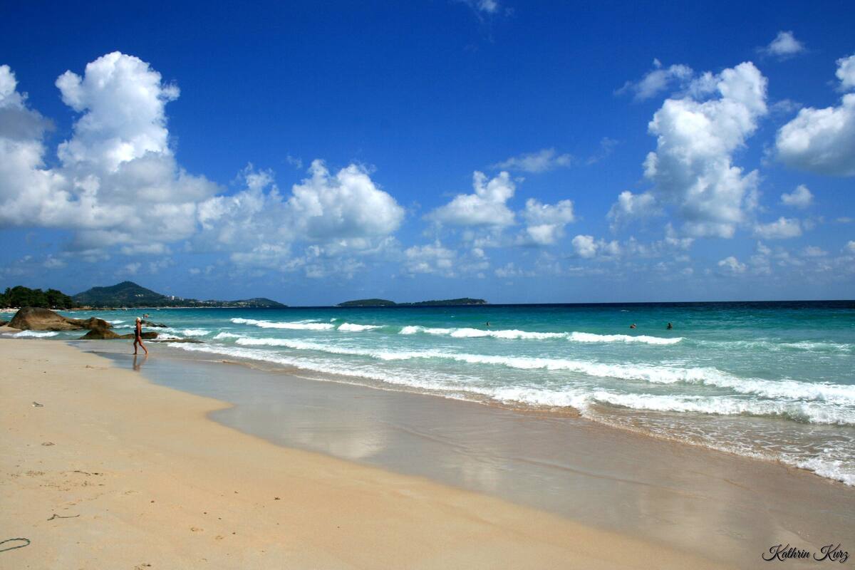 Una playa con cielos despejados y olas, una persona caminando, e islas a lo lejos.