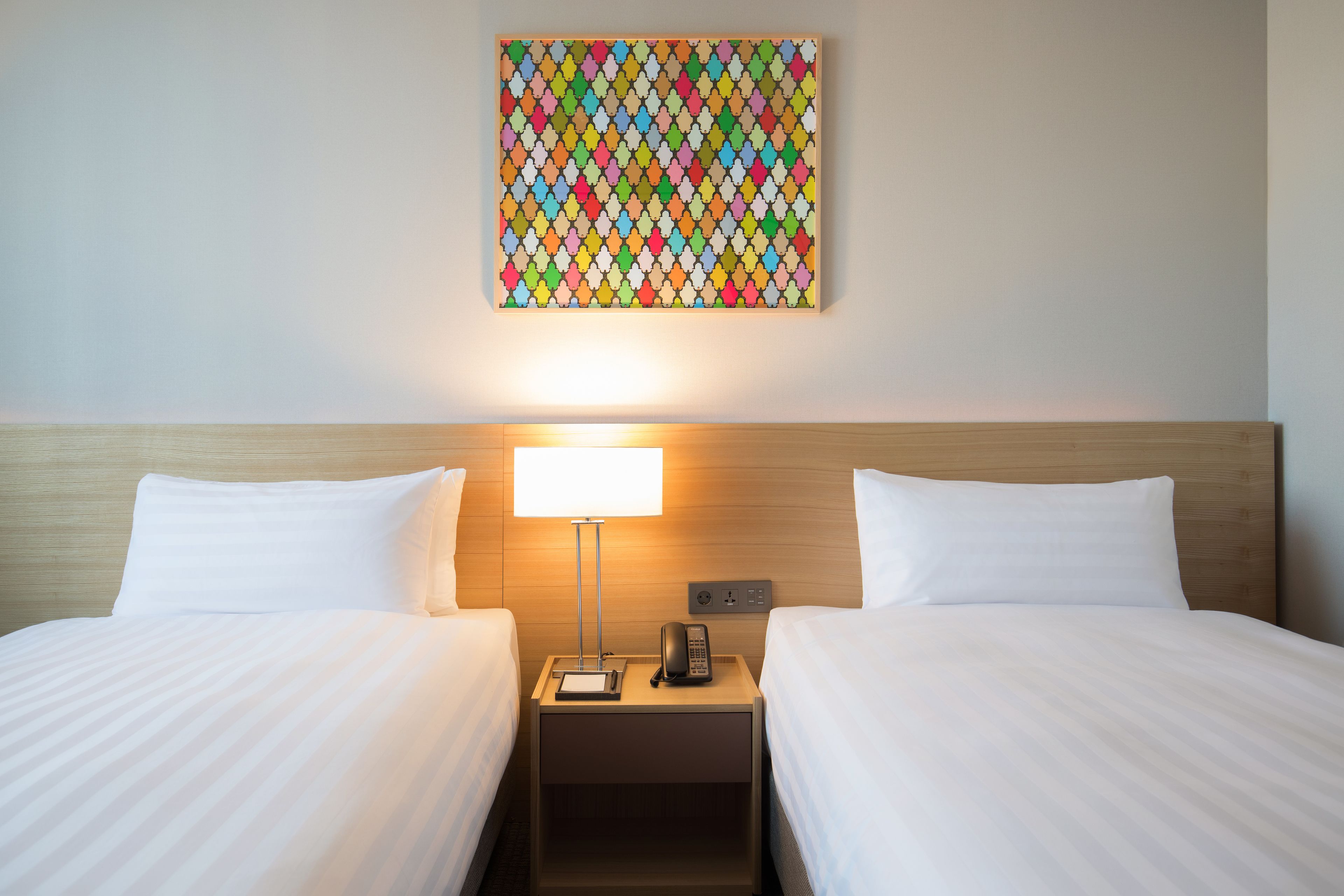 Habitación de hotel con dos camas, una mesita de noche con lámpara y una obra de arte colorida en la pared.