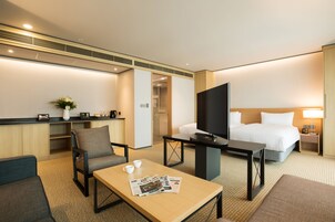 A hotel room with a bed, a TV, a desk with a chair, a coffee table with newspapers, and a small plant.