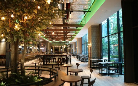 A spacious indoor dining area with multiple tables and chairs, a large tree, and large windows allowing natural light.