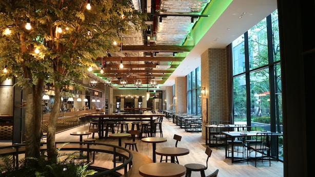 A spacious indoor dining area with multiple tables and chairs, a large tree, and large windows allowing natural light.