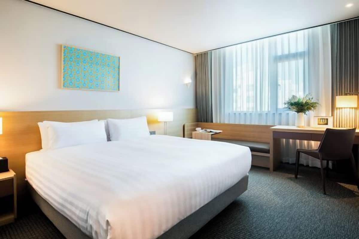 Une chambre d’hôtel avec un grand lit, un bureau, une chaise, une table de chevet, une lampe, une fenêtre avec des rideaux et un tableau au mur.