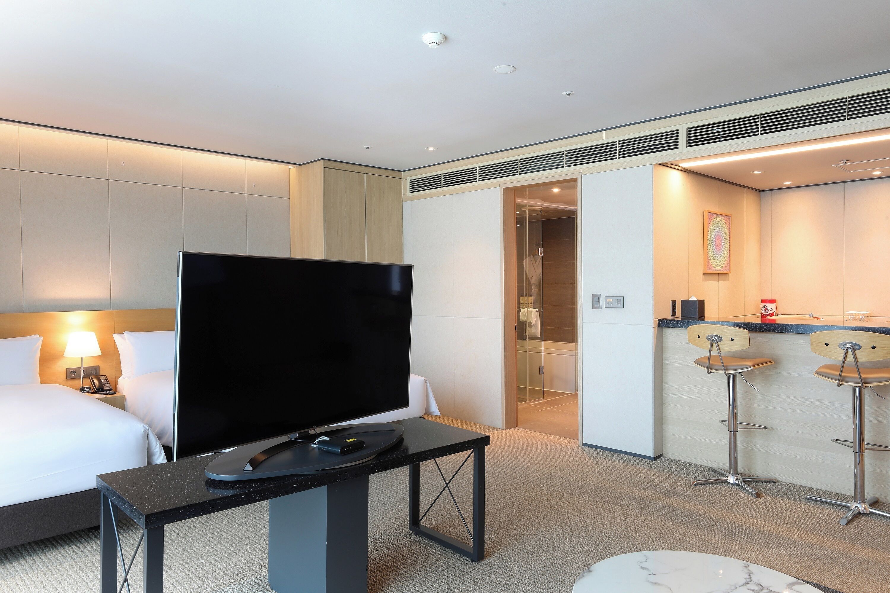 A hotel room with a bed, a flat-screen TV, a small table, a coffee machine, and a bathroom visible through an open door.