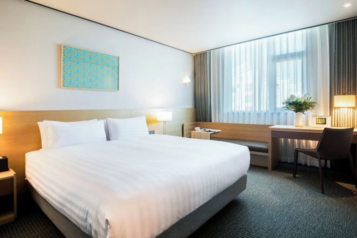 A hotel room with a large bed, a desk, a chair, a nightstand, a lamp, a window with curtains, and a painting on the wall.
