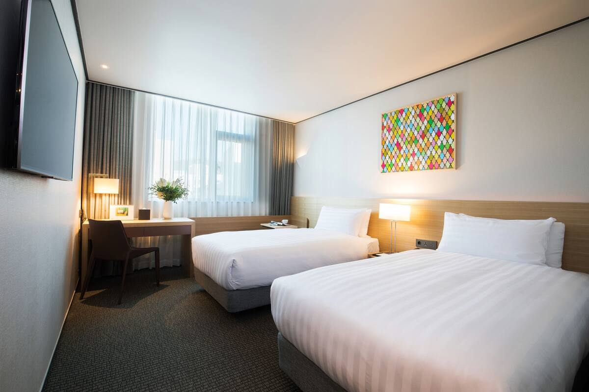 Une chambre d’hôtel avec deux lits, un bureau, une chaise, une télévision et une œuvre d’art colorée au mur.